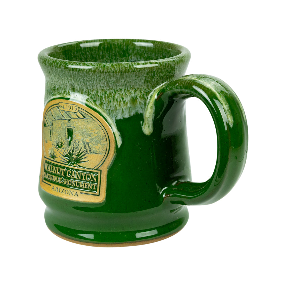 A green glazed ceramic mug with a handle, featuring a gold-colored 'Walnut Canyon Heritage Monument Arizona' label on the side, set against a plain white background.