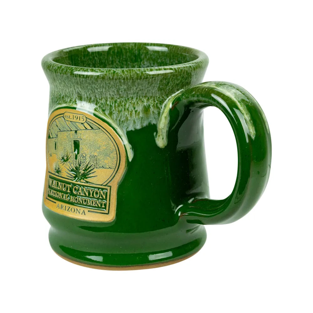 A green glazed ceramic mug with a handle, featuring a gold-colored 'Walnut Canyon Heritage Monument Arizona' label on the side, set against a plain white background.