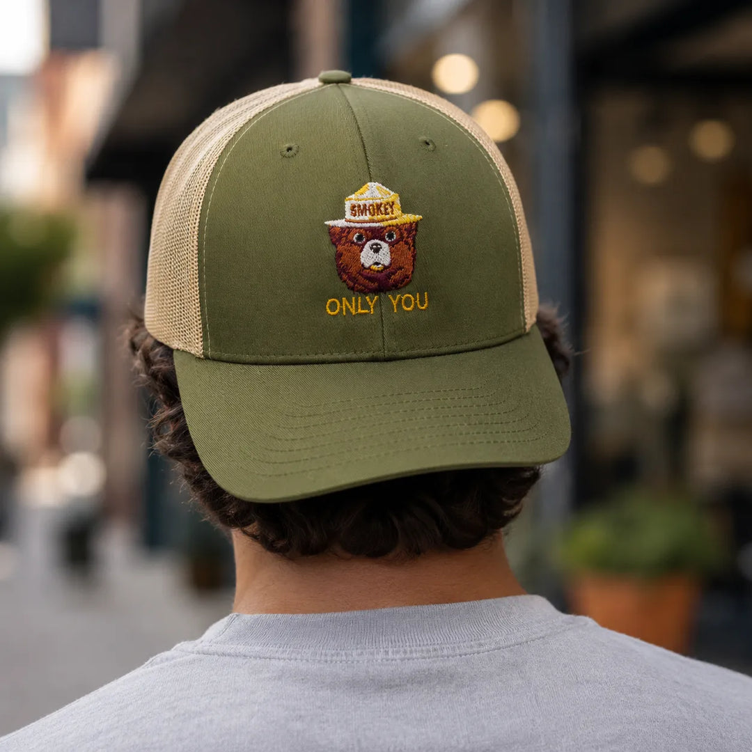 A man viewed from behind wearing an olive green and beige trucker hat with a bear graphic and the text &