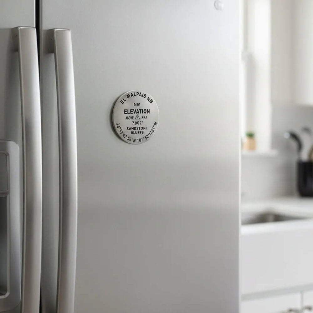 A round metallic elevation marker plaque for El Malpais NM, showing engraved text including 'ELEVATION', '7,002 feet', and geographic coordinates on a stainless steel refrigerator.