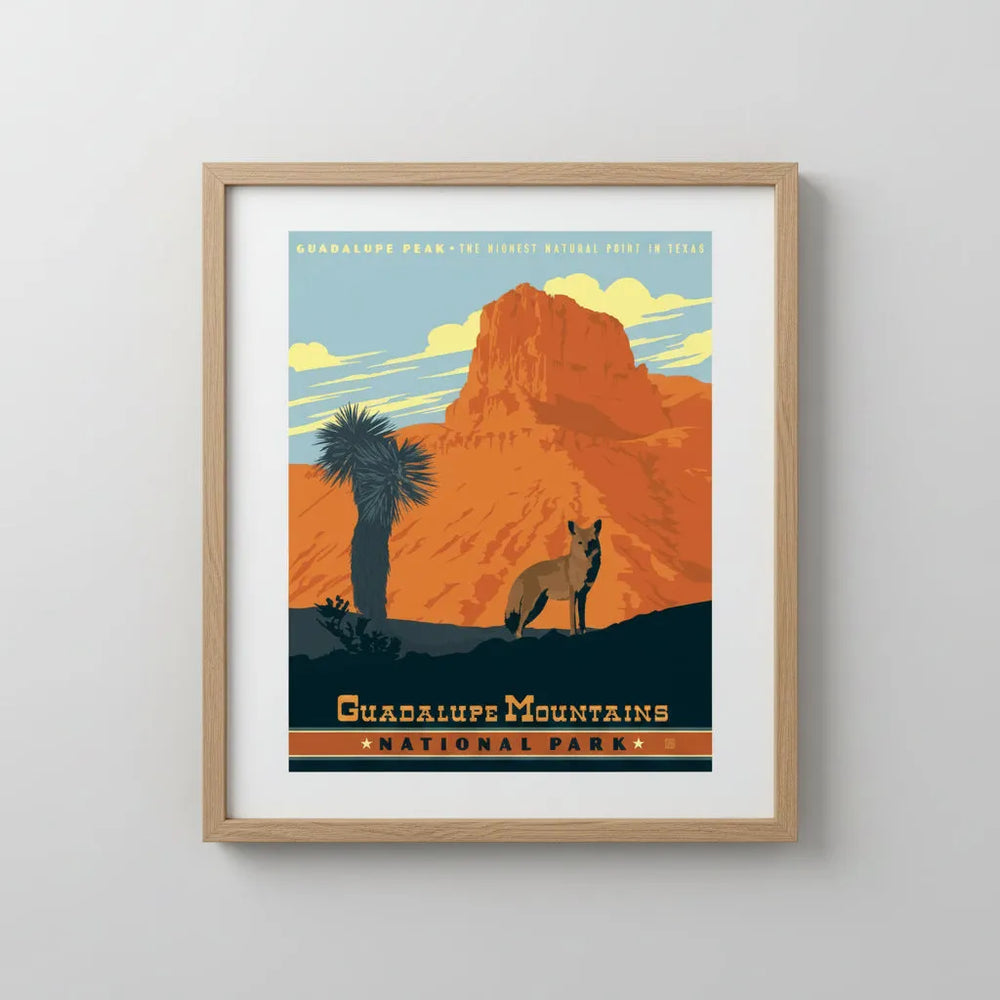 A vintage-style travel poster for Guadalupe Mountains National Park, featuring an orange rock formation, a blue sky, a palm tree, and a coyote, mounted and framed, hanging on a wall.