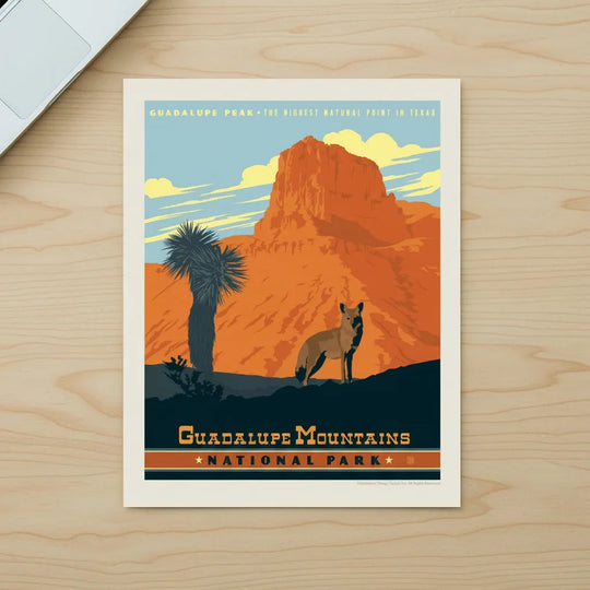 A vintage-style travel poster for Guadalupe Mountains National Park, featuring an orange rock formation, a blue sky, a palm tree, and a coyote, lying flat on a desk.