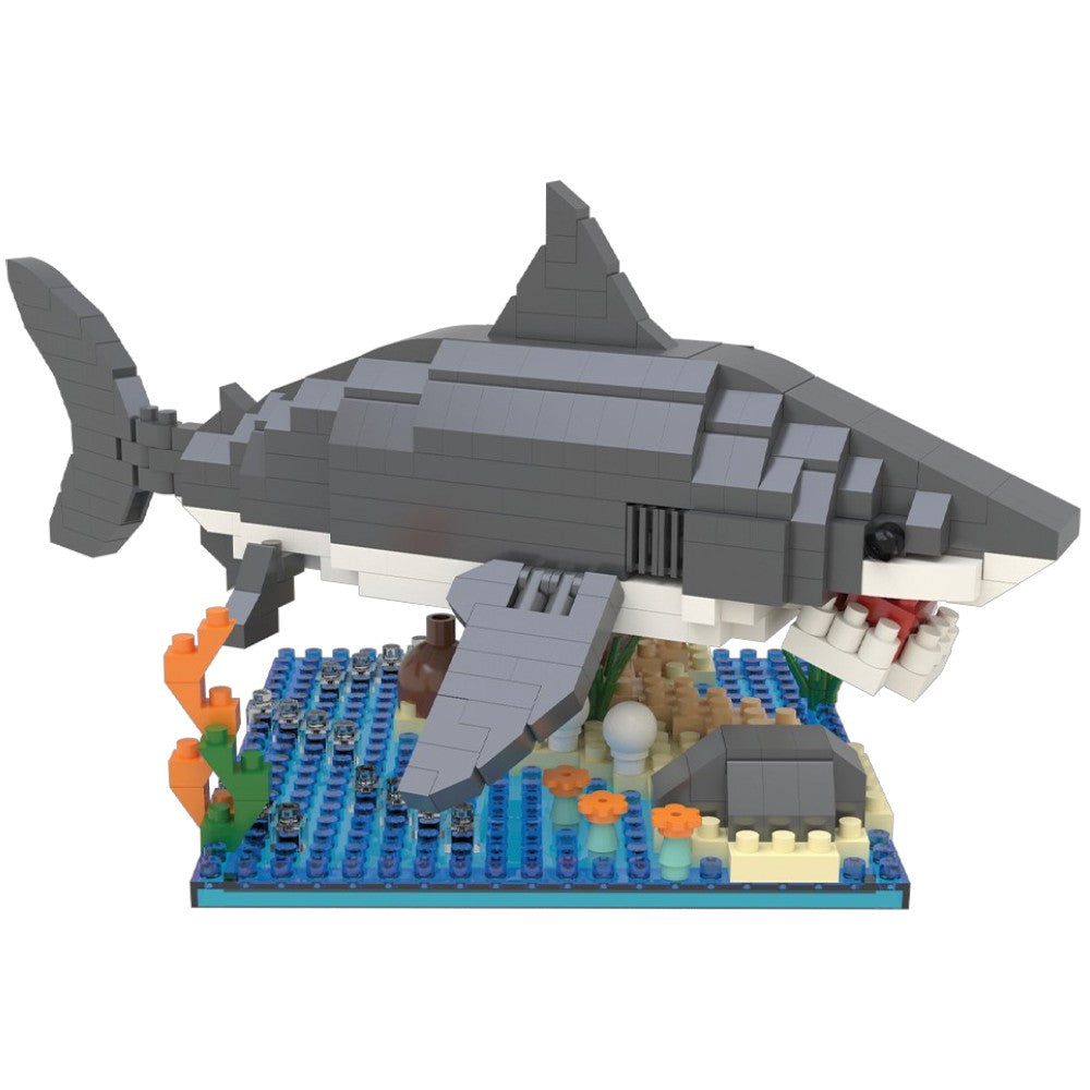 A block-built model of a shark swimming over an underwater ocean floor scene, constructed from interlocking toy bricks. The shark is primarily gray and white, with a blue ocean base featuring coral in orange, green, and brown.