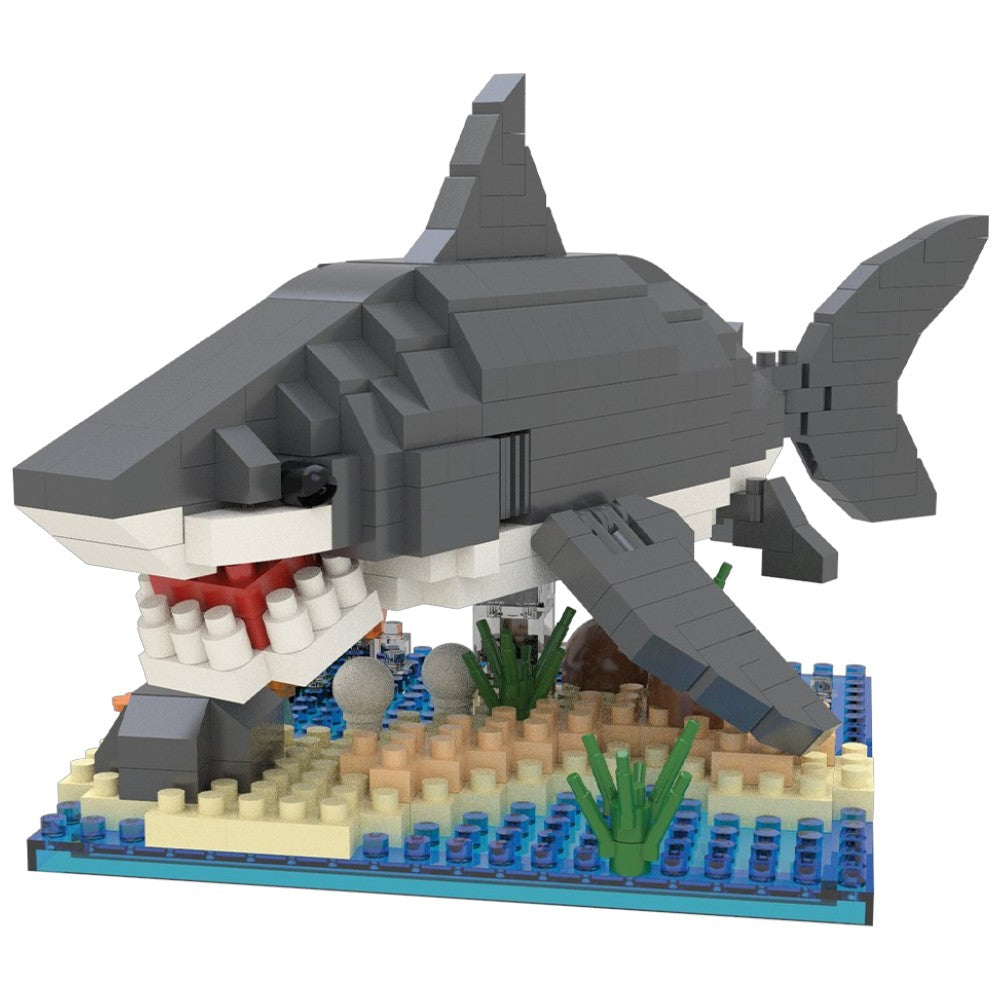 A toy building-block shark model with gray, white, and red details, emerging from an underwater sand and blue-water base with green seaweed.