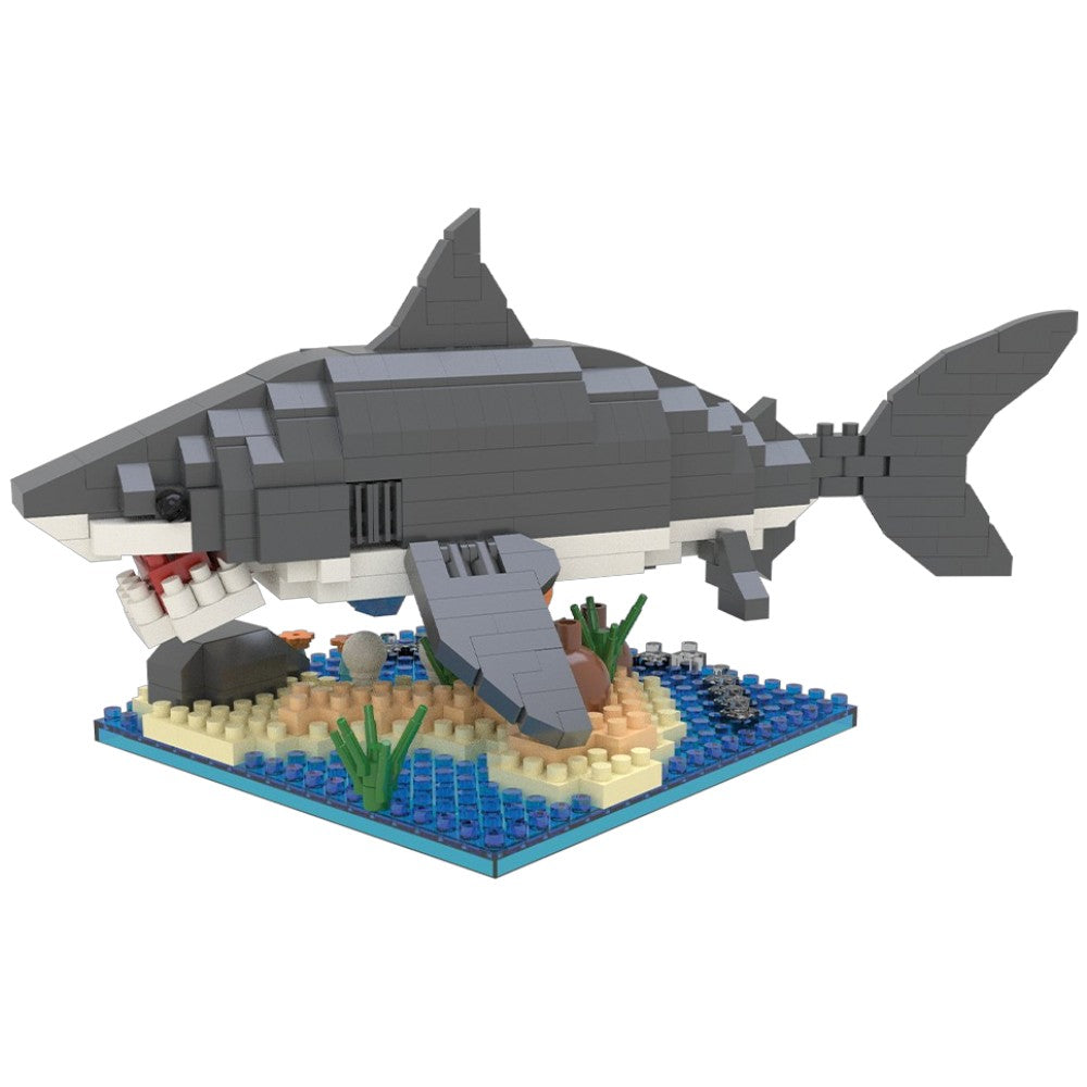 A gray and white toy shark model built from small interlocking blocks, positioned above a blue ocean floor base with sand, rocks, and green seaweed, on a plain white background.