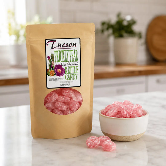 A stand-up kraft pouch of Tucson Prickly Pear Old Fashioned Kettle Candy stands on a marble countertop with a small white bowl and loose pink candy in front, set in a softly blurred kitchen background.