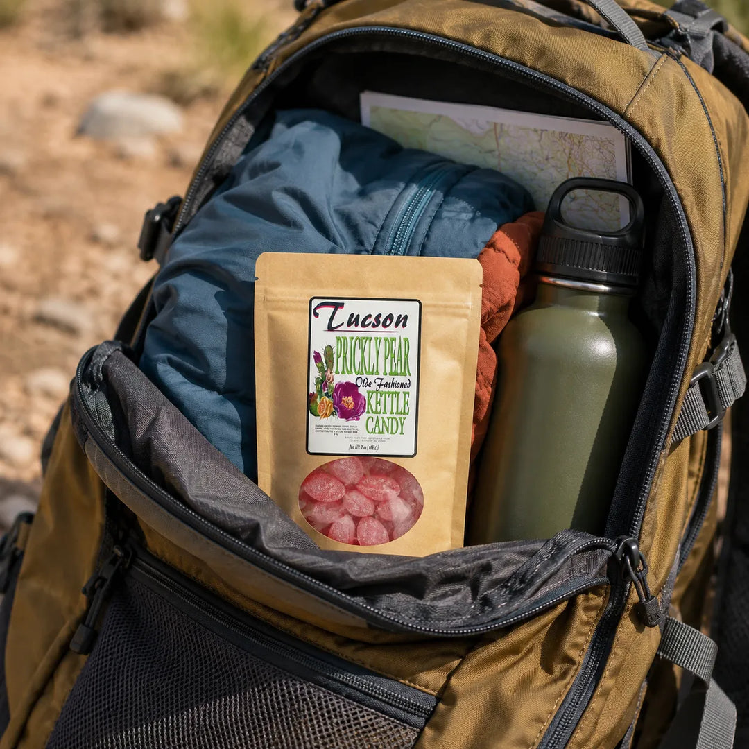 An open tan backpack on a trail shows its contents, including a pouch of Tucson Hickory Bear natural candy, a blue jacket, an olive green water bottle, and a map.