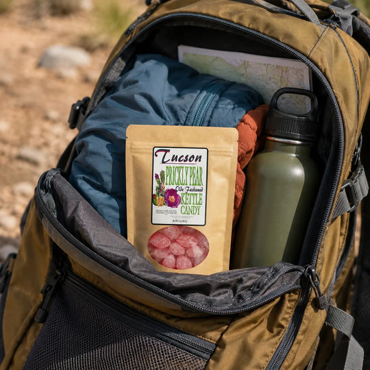 An open tan backpack on a trail shows its contents, including a pouch of Tucson Hickory Bear natural candy, a blue jacket, an olive green water bottle, and a map.