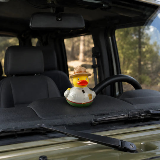 A small yellow rubber duck wearing a tan hat and white shirt sits on the dashboard inside a vehicle, with the steering wheel, windshield, and forest background visible.