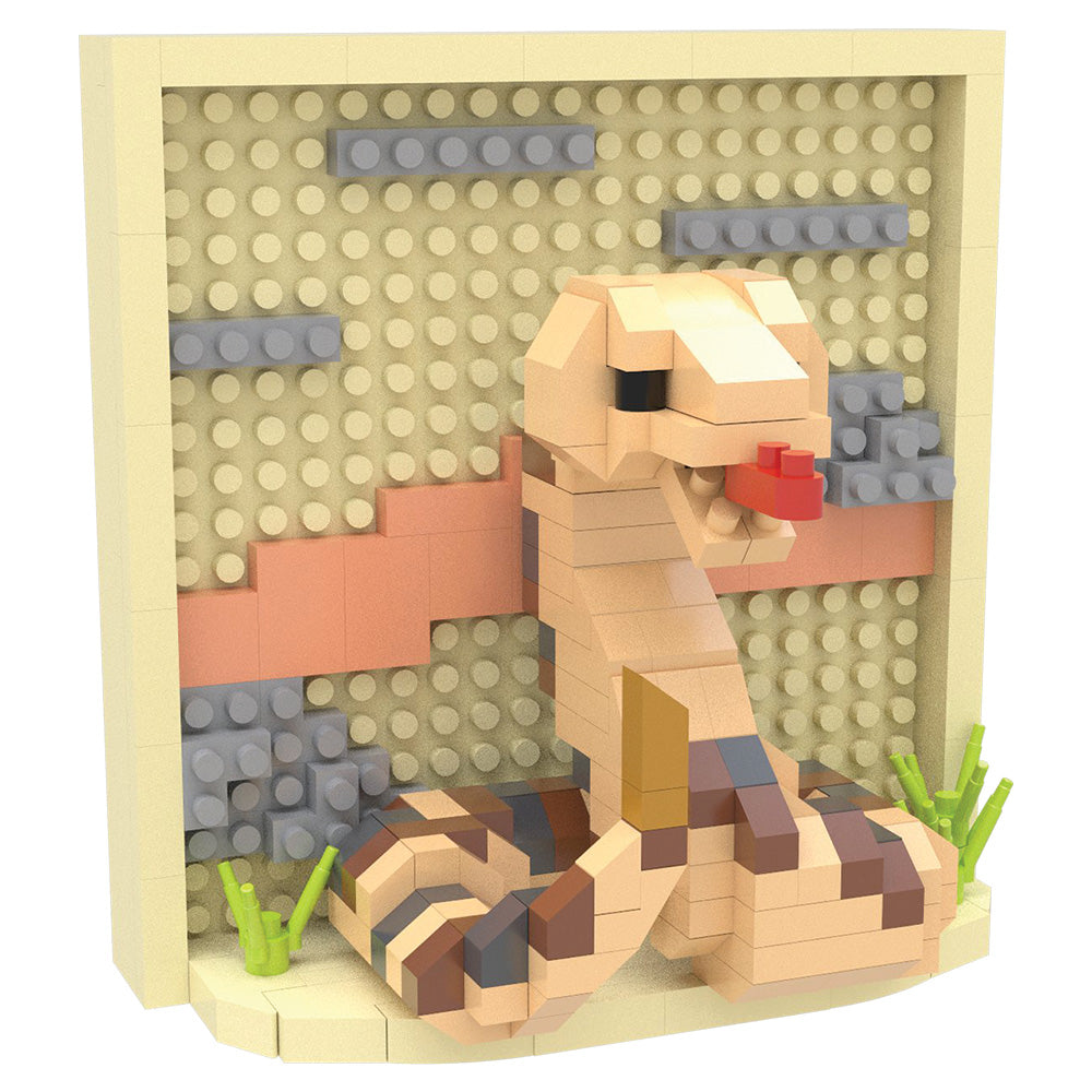 A toy building-block snake model posed inside a beige display frame with a textured beige background, gray rock details, a pink stripe, green grass pieces, and a small red tongue.