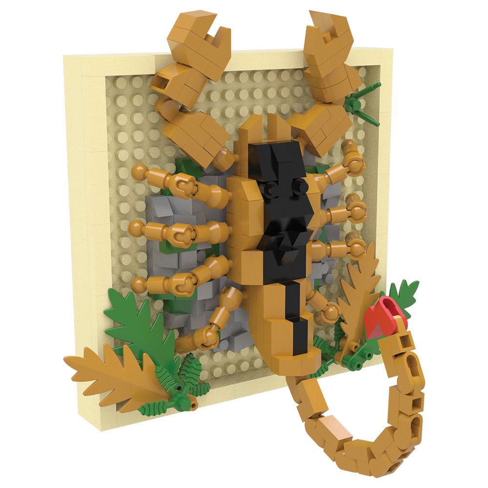 A toy building-block model of a scorpion mounted on a beige display board, with tan, black, gray, green, and small red details, shown on a plain white background.