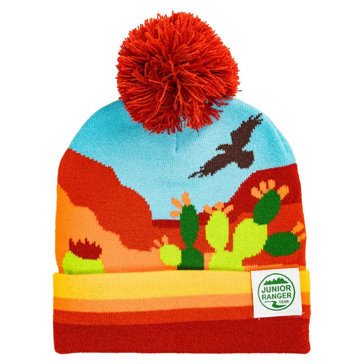 A colorful Southwest Junior Ranger Beanie with a pom-pom, featuring a desert landscape with mesas, cacti, and a bird of prey.
