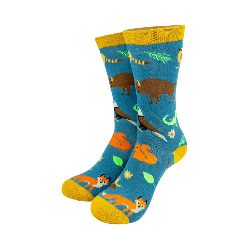 A pair of crew socks with a colorful nature-themed pattern featuring brown bison, orange foxes, green leaves, and a penguin on a blue background with yellow cuffs, heels, and toes.