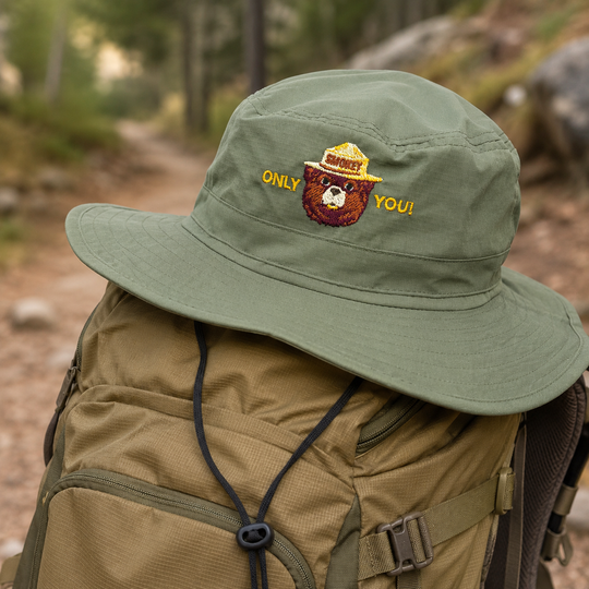 An olive green wide-brim bucket hat with embroidered text and a bear graphic, resting on top of a tan hiking backpack on a forest trail.