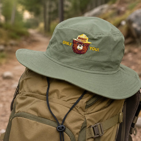 An olive green wide-brim bucket hat with embroidered text and a bear graphic, resting on top of a tan hiking backpack on a forest trail.