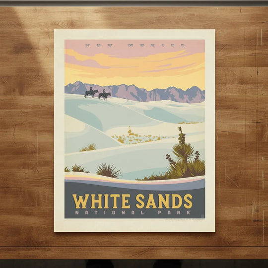 A vintage-style travel poster for White Sands National Park in New Mexico, showing pale blue-white sand dunes, distant purple mountains, and a warm yellow-orange sky, lying flat on a drafting table.