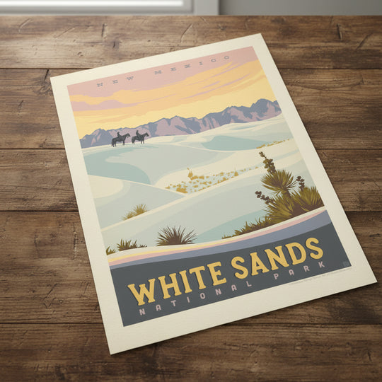 A vintage-style travel poster for White Sands National Park in New Mexico, showing pale blue-white sand dunes, distant purple mountains, and a warm yellow-orange sky, lying flat on a rustic table.