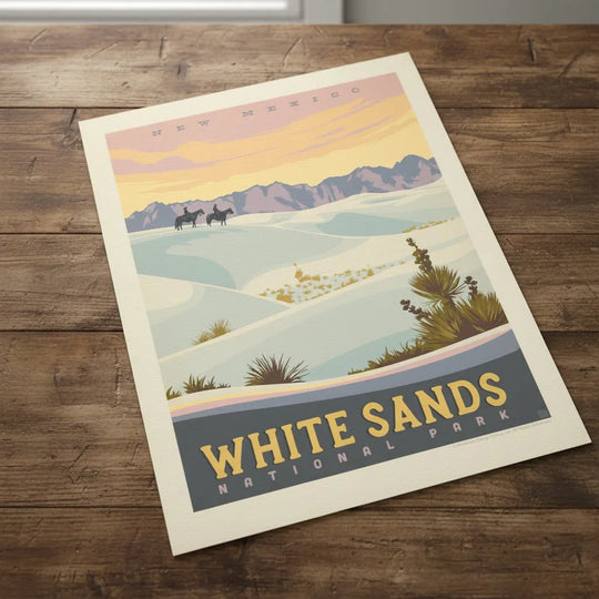 A vintage-style travel poster for White Sands National Park in New Mexico, showing pale blue-white sand dunes, distant purple mountains, and a warm yellow-orange sky, lying flat on a rustic table.