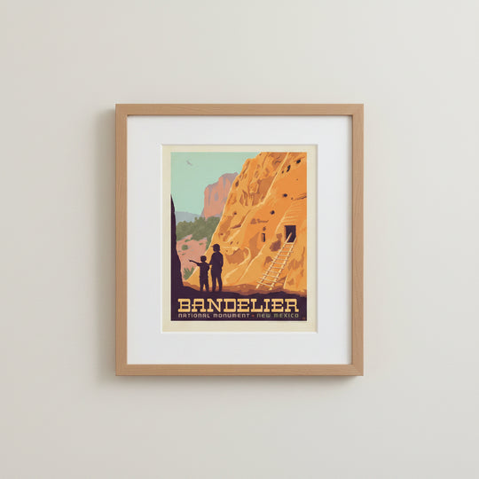 A vintage-style travel poster for Bandelier National Monument in New Mexico, showing a large orange rock cliff with a wooden ladder, silhouetted figures at the base, framed and mounted on a wall.