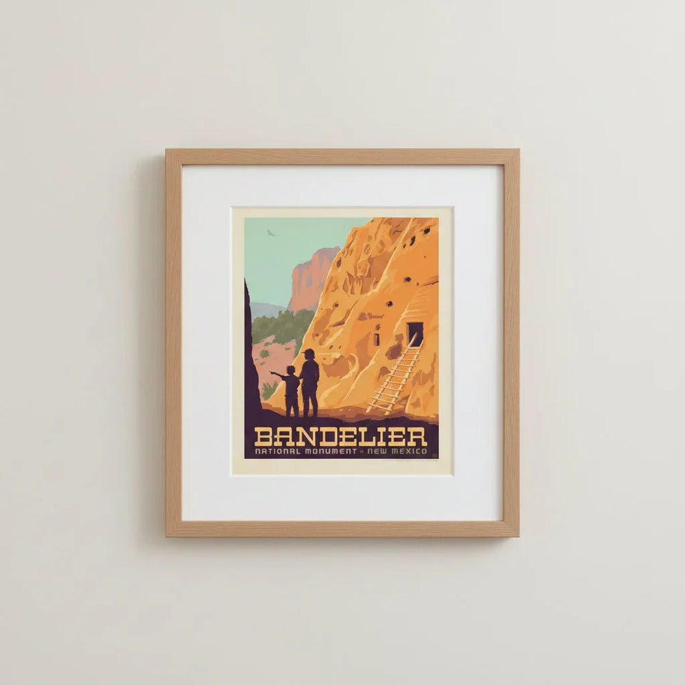 A vintage-style travel poster for Bandelier National Monument in New Mexico, showing a large orange rock cliff with a wooden ladder, silhouetted figures at the base, framed and mounted on a wall.