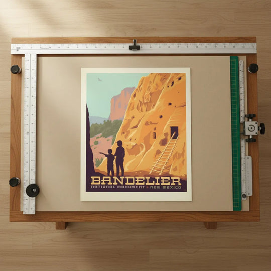 A vintage-style travel poster for Bandelier National Monument in New Mexico, showing a large orange rock cliff with a wooden ladder, silhouetted figures at the base, placed on a drafting table.