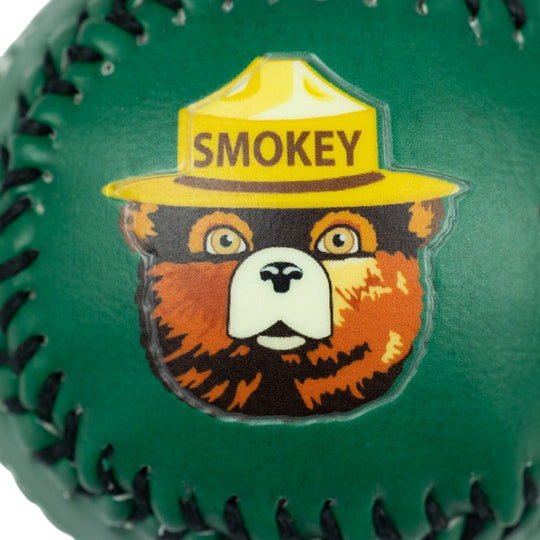 A close-up of a green baseball with black stitching and a large printed Smokey Bear logo on the front. The bear wears a yellow hat with the word &