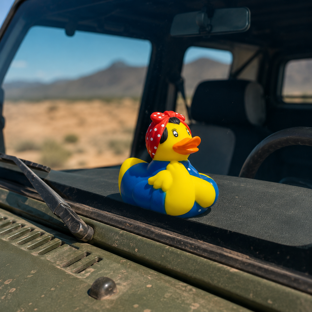 A yellow rubber duck wearing a red polka-dot bandana and blue overalls sits on the dashboard of an olive green vehicle, with a blurred desert landscape and mountains visible through the windshield.