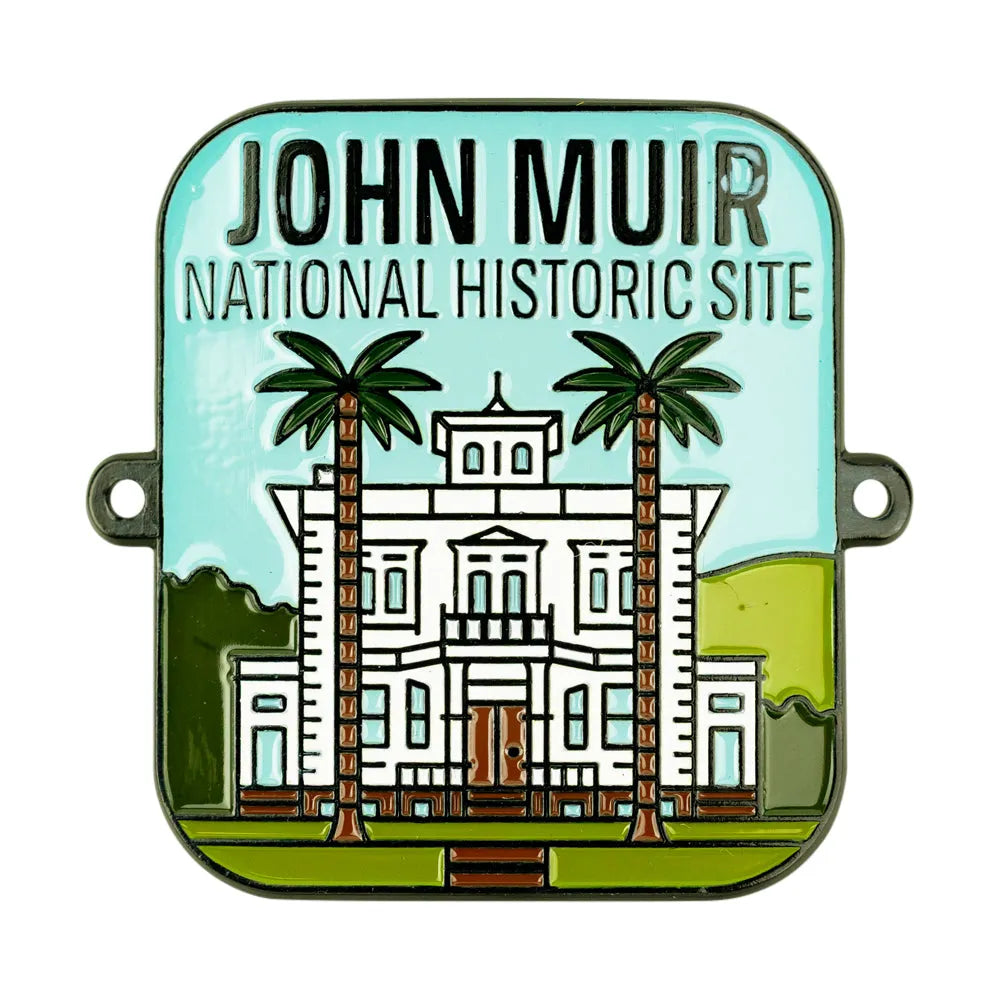 A square enamel hiking stick medallion featuring an illustration of the John Muir National Historic Site building with two palm trees, set against a light blue sky background with the text 'JOHN MUIR NATIONAL HISTORIC SITE' at the top.