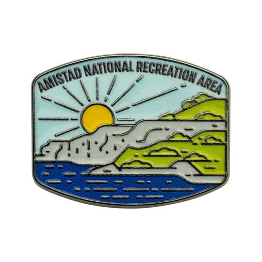 An enamel pin depicting the Amistad National Recreation Area, featuring a yellow sun over blue water and green hills under a light blue sky, with the text &