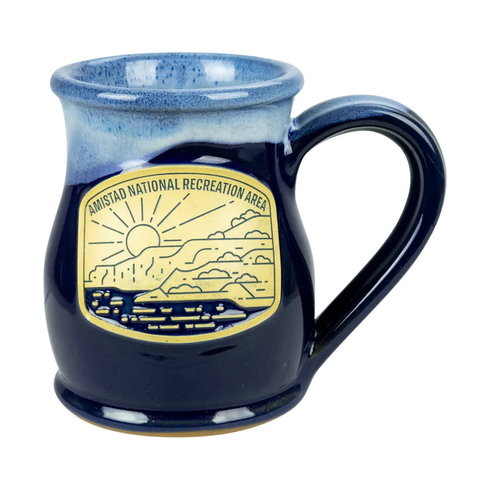 A dark blue glazed ceramic mug with a handle, featuring a gold-colored Amistad National Recreation Area emblem on the front.