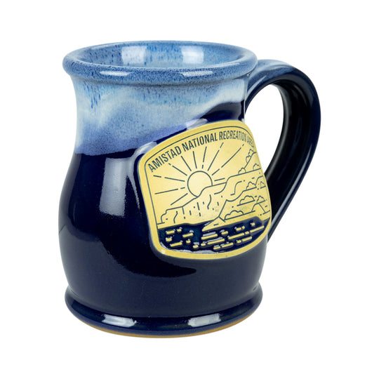 A ceramic mug with a blue gradient glaze, featuring a gold-colored metal badge on the side that reads &