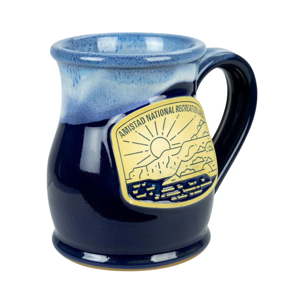 A ceramic mug with a blue gradient glaze, featuring a gold-colored metal badge on the side that reads &