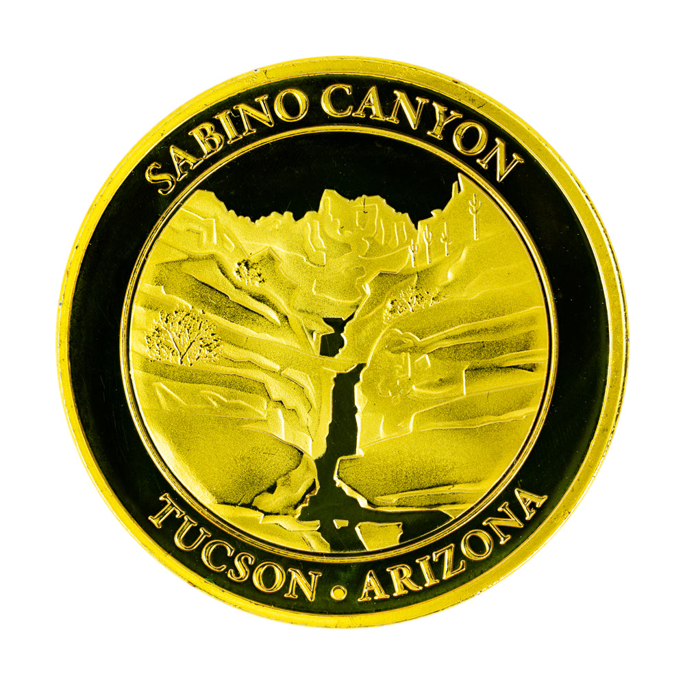 A round gold-colored commemorative coin for Sabino Canyon in Tucson, Arizona, featuring an embossed view of seven falls on a white background.