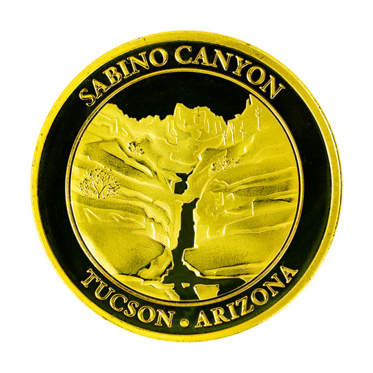 A round gold-colored commemorative coin for Sabino Canyon in Tucson, Arizona, featuring an embossed view of seven falls on a white background.