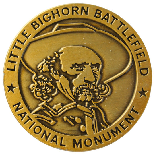 Gold-colored emblem of Little Bighorn Battlefield National Monument with a profiled face and floral design.