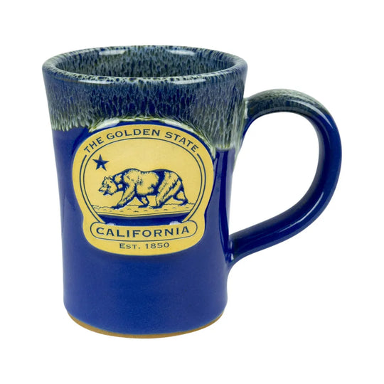 A ceramic coffee mug with a deep blue body, a textured dark blue and green glazed rim, and a yellow circular California-themed emblem on the front featuring a grizzly bear and the text &