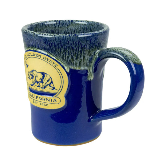 A ceramic coffee mug with a deep blue glazed finish, featuring a green and blue speckled glaze around the rim and handle. The front is decorated with a yellow circular &