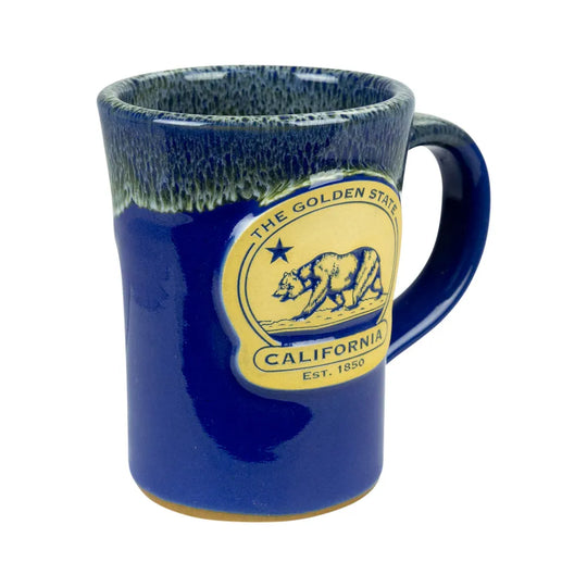 A ceramic coffee mug with a deep blue body, a textured dark blue and green glazed rim, and a yellow circular California-themed emblem on the side.