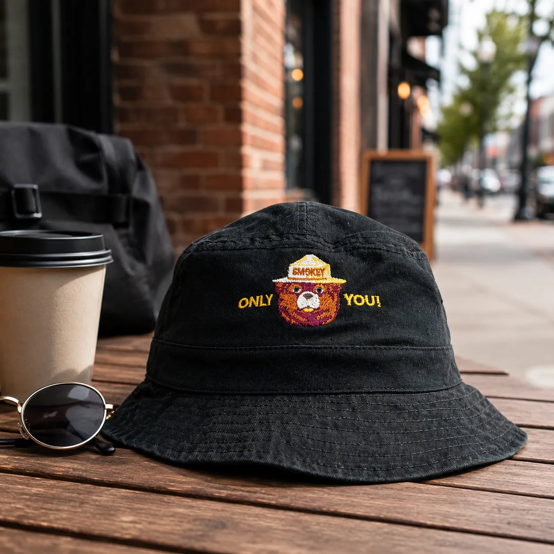 A black bucket hat with an embroidered design sits on a wooden table outdoors. The embroidery features a yellow and orange design with the words &