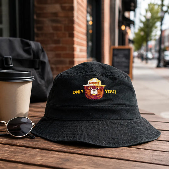 A black bucket hat with an embroidered design sits on a wooden table outdoors. The embroidery features a yellow and orange design with the words &