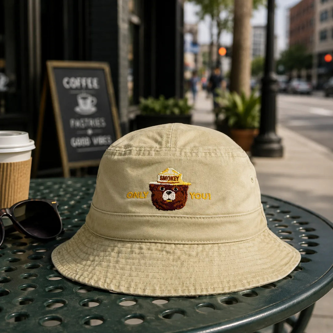 A beige bucket hat with an embroidered brown bear wearing a yellow hat and the words &