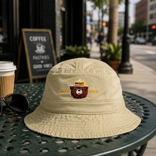A beige bucket hat with an embroidered brown bear wearing a yellow hat and the words &