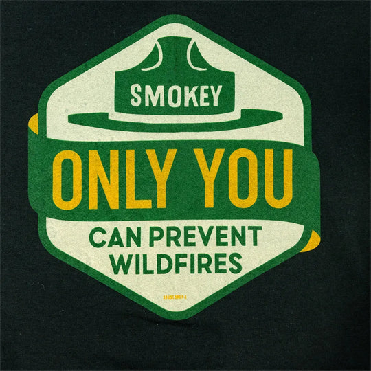 A close-up of a dark charcoal fabric featuring a large graphic print. The graphic is a hexagonal badge with a green border, cream background, and bold yellow and green text reading &