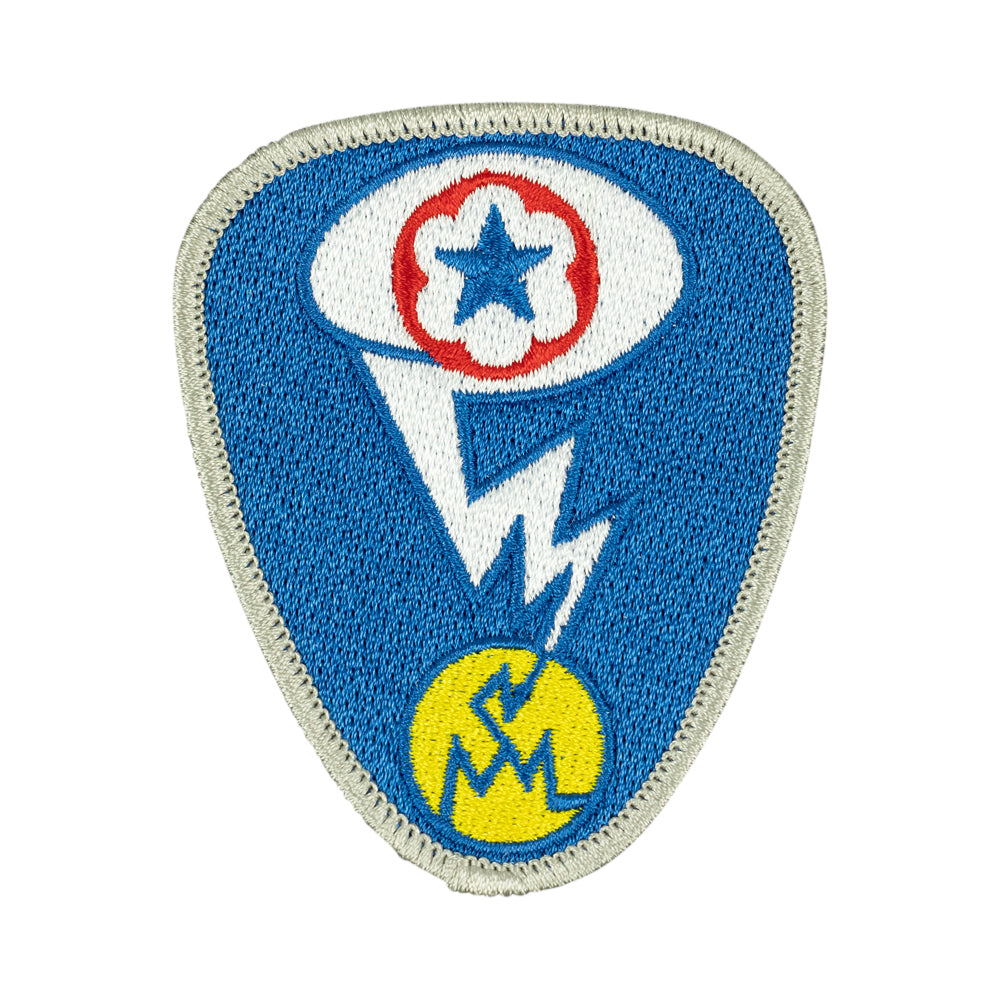 An embroidered shield-shaped patch featuring a blue field with a white border, a white eye motif containing a red circle and blue star, a white lightning bolt, and a yellow circle near the bottom with a blue symbol.