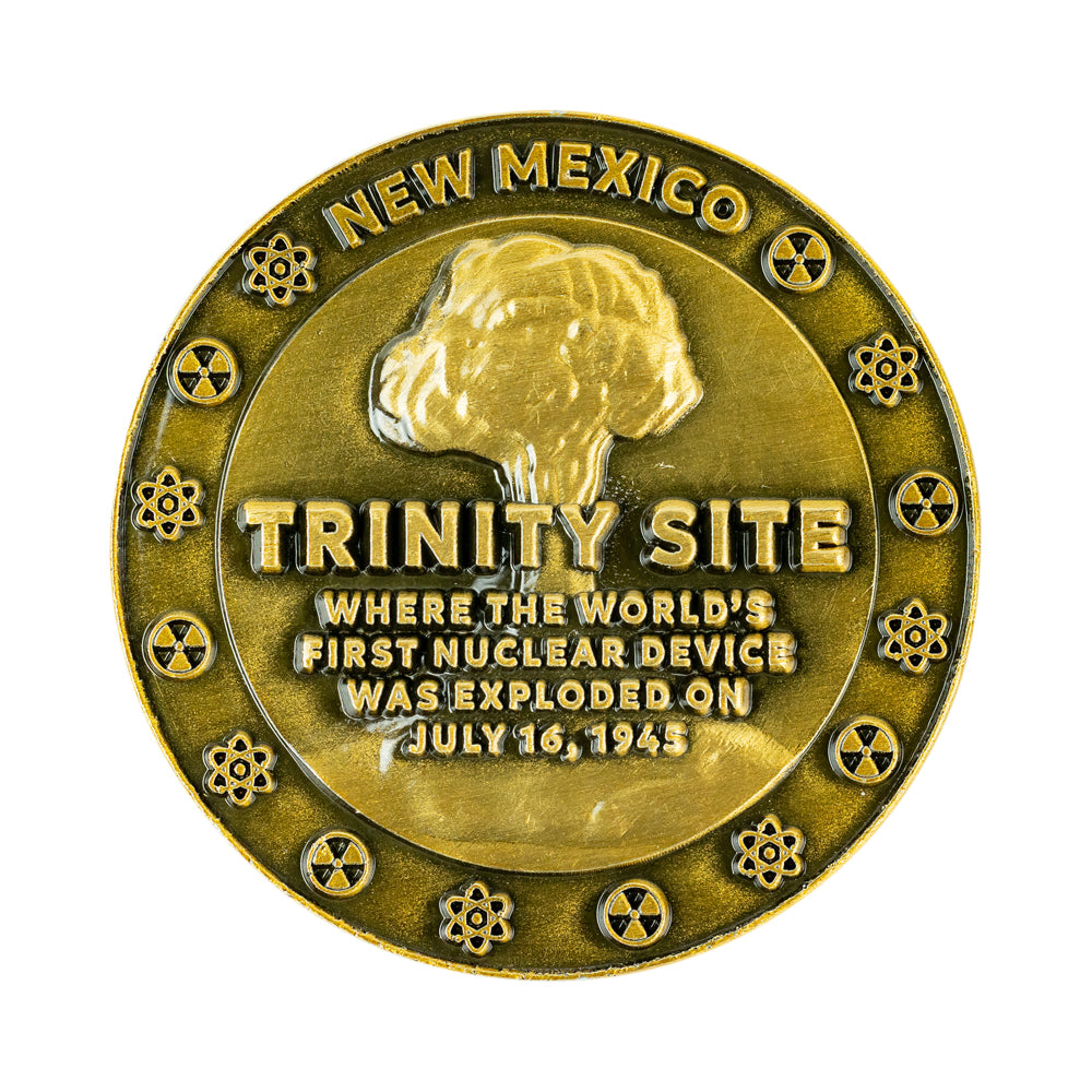 A round metallic commemorative coin featuring a Trinity Site nuclear explosion design with the text &