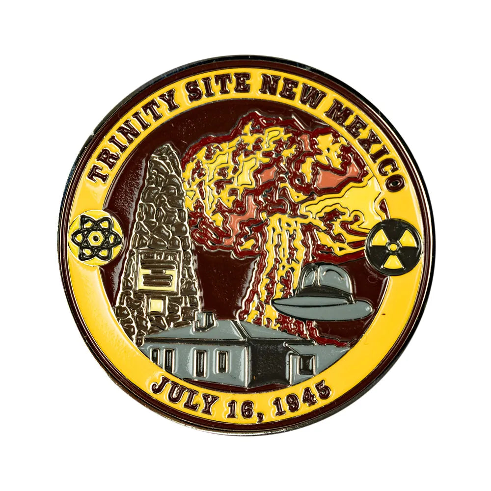 A circular magnet reading 'TRINITY SITE NEW MEXICO' and 'JULY 16, 1945,' with an explosion, building, house, and radiation symbol in gold, red, gray, black, and white enamel.