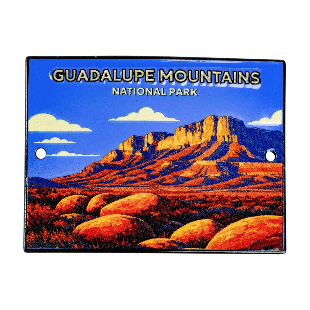 A rectangular metal medallon featuring a colorful illustration of Guadalupe Mountains National Park, with a blue sky, white clouds, and orange-red rock formations. The text &