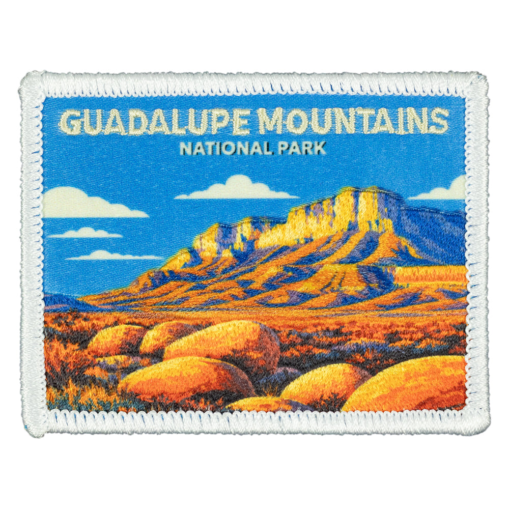 An embroidered patch featuring a landscape illustration of Guadalupe Mountains National Park, with a blue sky, white clouds, and orange-brown mountains and rocks, bordered by a white stitched edge.