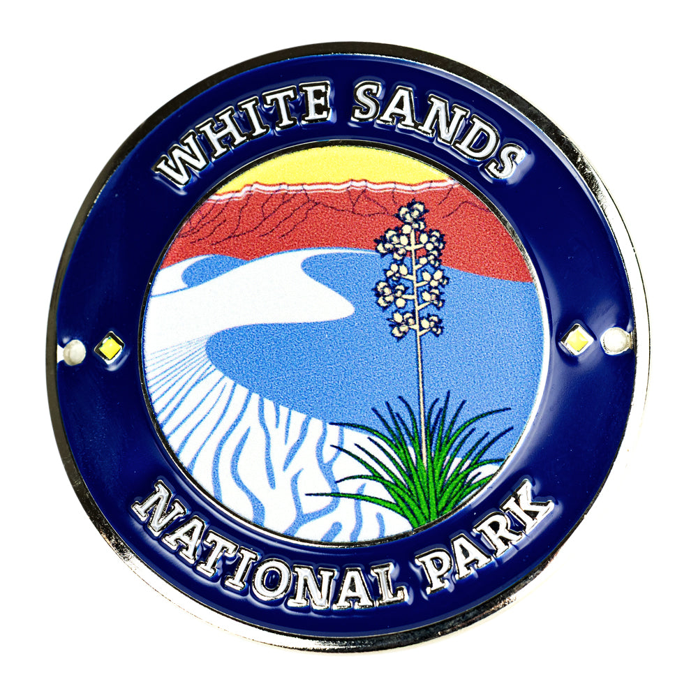 A circular enamel-style medallionreading “WHITE SANDS NATIONAL PARK,” with a blue border and a central scenic illustration of white dunes, red mountains, and green grass on a white background.