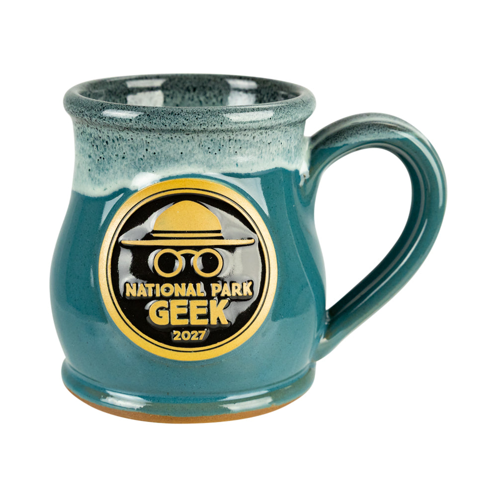 A teal ceramic mug with a speckled glaze and a gold and black National Park Geek 2027 commemorative patch on the front, set against a plain white background.